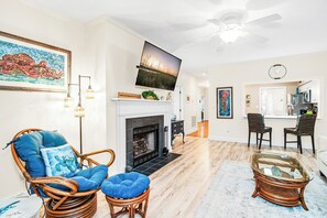 Ein Wohnzimmer mit Kamin, einem darüber montierten Fernseher, Korbmöbeln und einer Küche im Hintergrund.