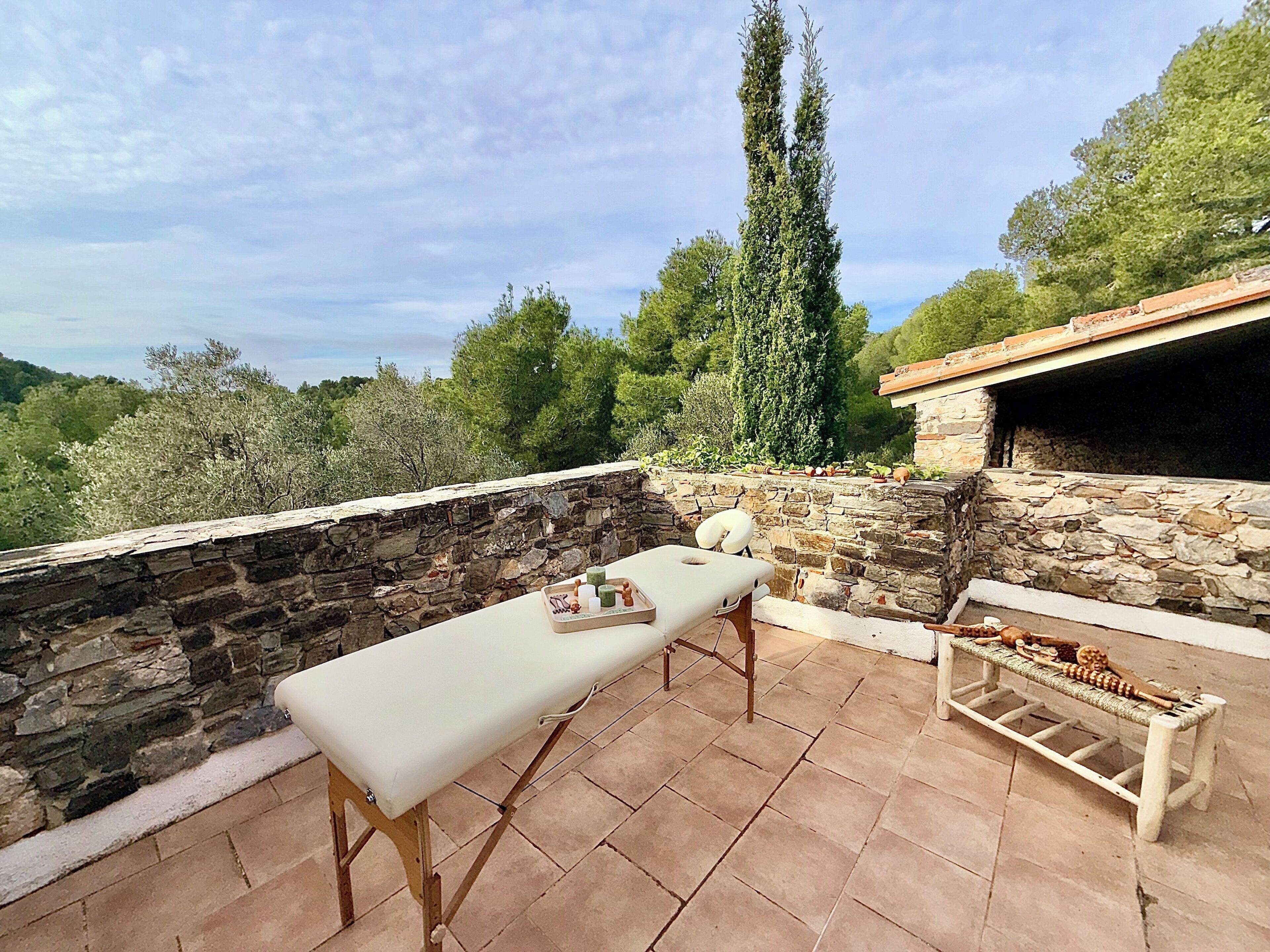 En terrasse med stenvægge, et bord og en bænk, omgivet af frodig vegetation og en blå himmel.