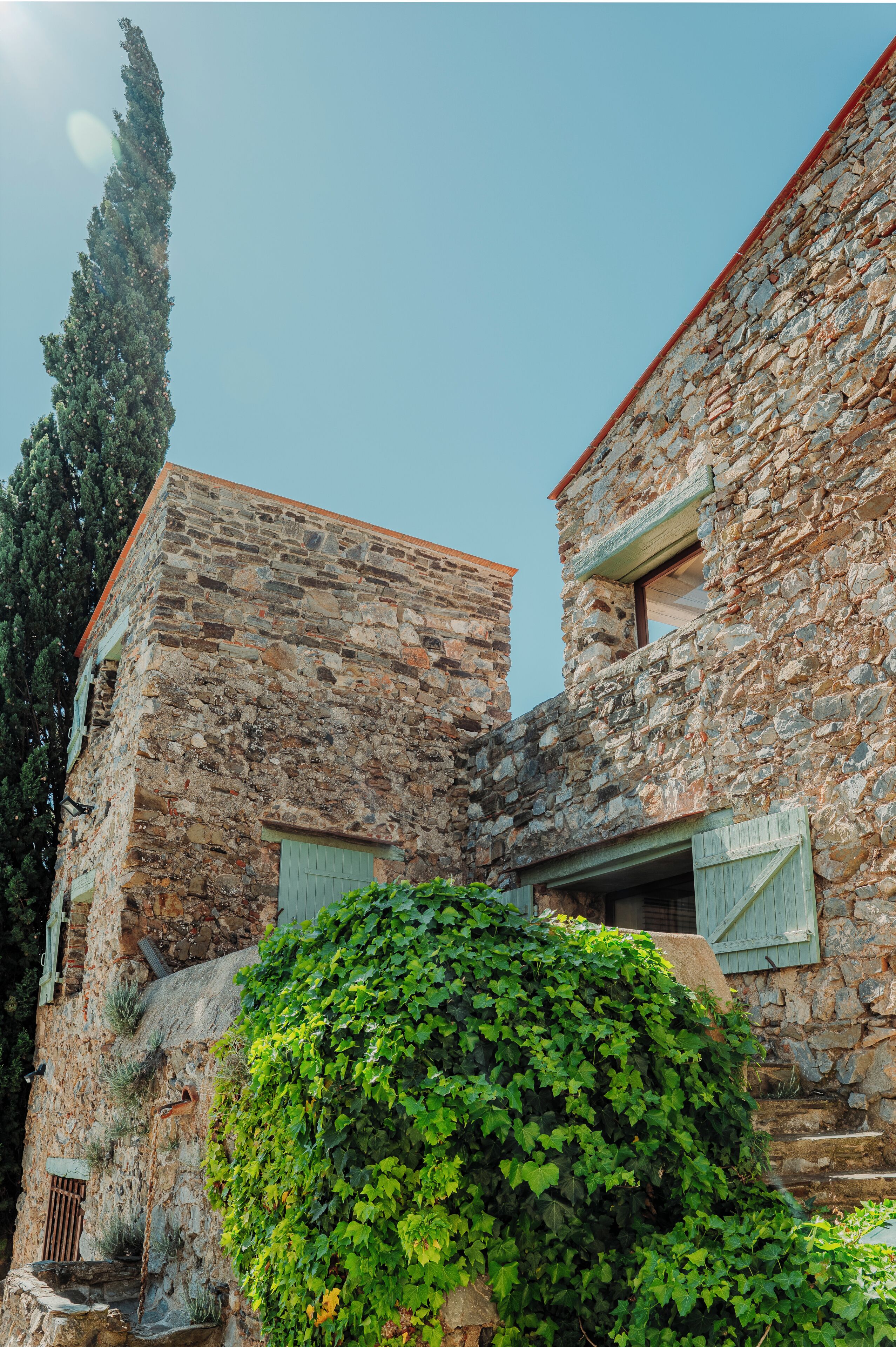 A stone building with a red roof, a small window with shutters, and a green bush in front.