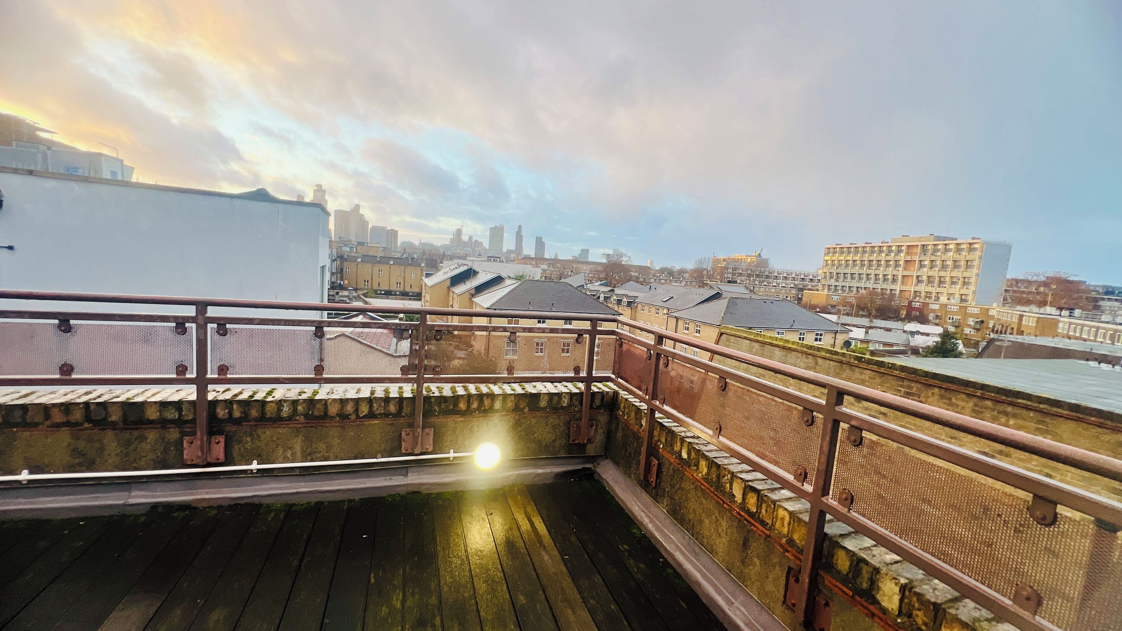 Una terraza en la azotea con barandilla de metal, con vistas a un paisaje urbano con edificios varios y un río.