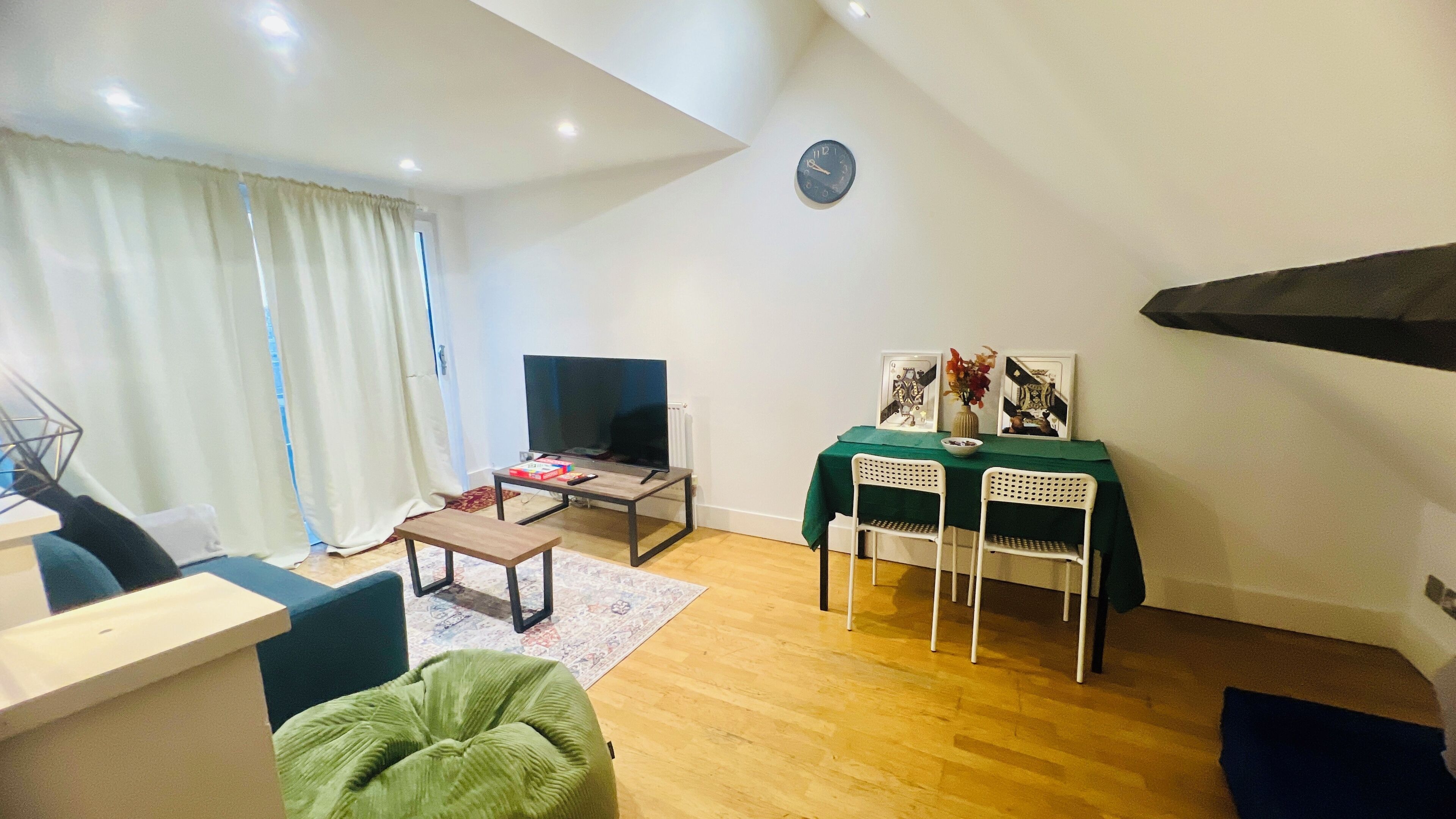 A living room with a TV, a small table with chairs, a green ottoman, and a clock on the wall.