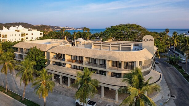 Un hotel de varios pisos con techo curvo, palmeras y vista al mar.