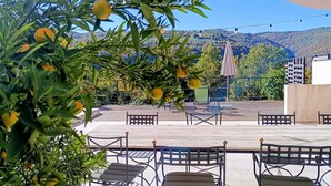 En udendørs terrasse med borde og stole, omgivet af grønt og et bjergrigt landskab.