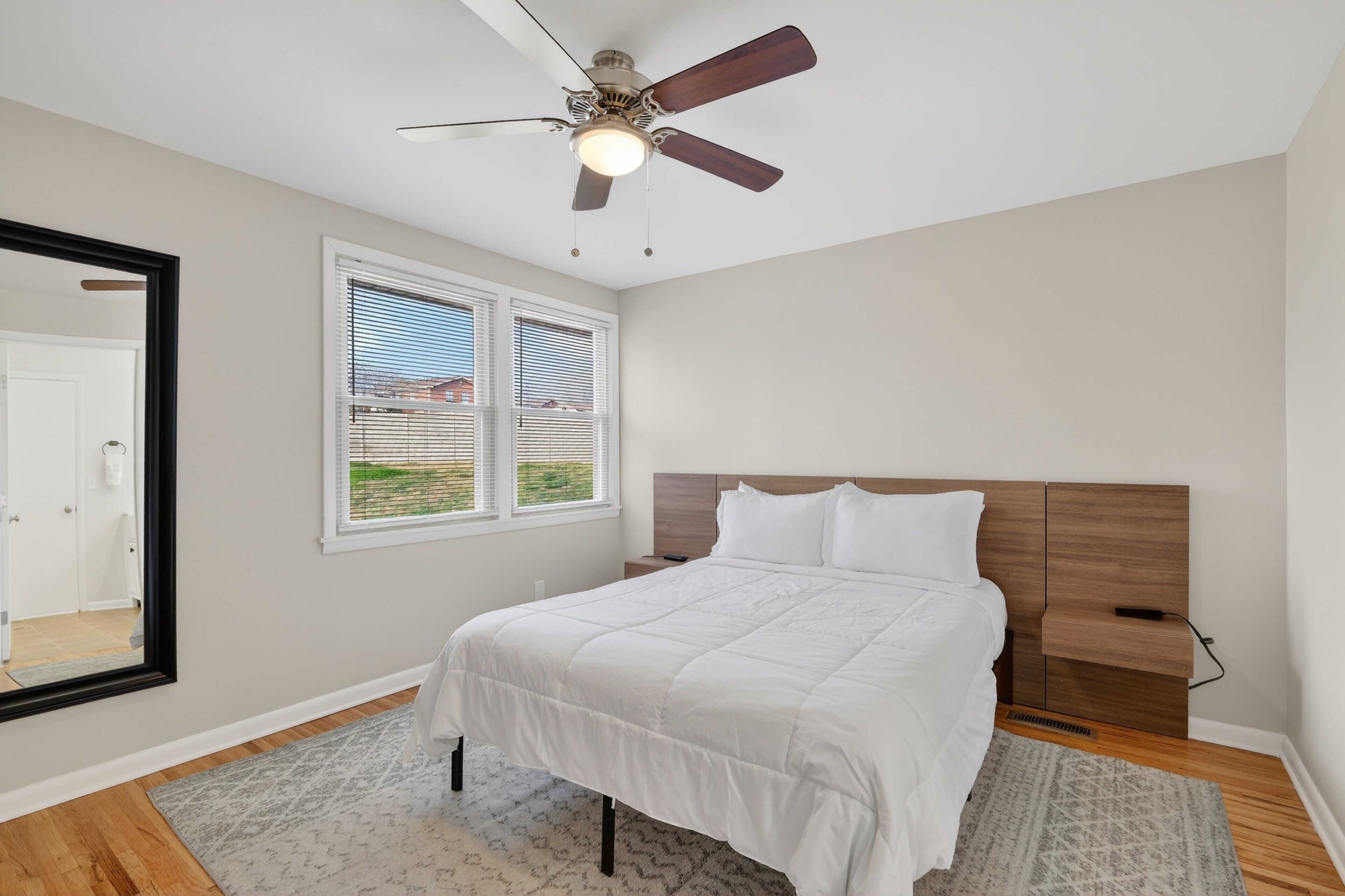 Une chambre à coucher comprenant un lit, un ventilateur de plafond, une fenêtre avec des stores, une table de chevet et un miroir.