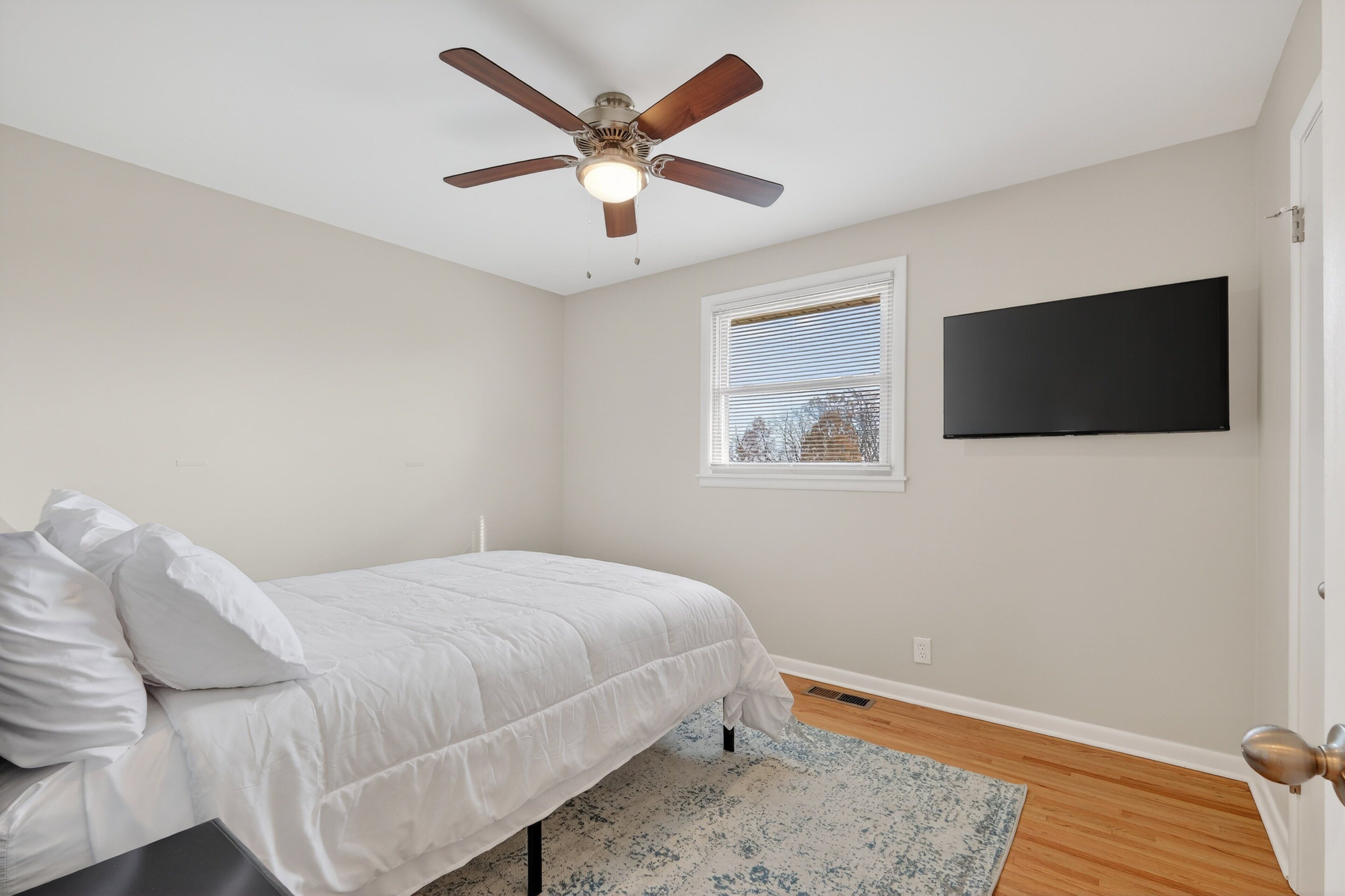 A bedroom with a bed, a ceiling fan, a window with blinds, and a flat-screen TV mounted on the wall.