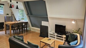 A modern living room with a wooden dining table, black leather sofa, and a flat-screen TV on a white stand. - Chic Family Hideaway in Durbuy (Durbuy)