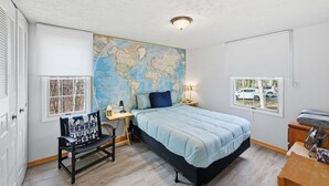 A bedroom with a large world map on the wall, a bed with a blue bedspread, a bench with a patterned cushion, and a window with a view of parked cars. - Manassas Comfort Cozy 3br Family Home 3 Bedroom Home (Manassas)