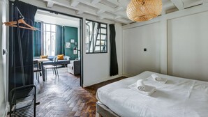 A hotel room with a large bed, a wooden floor, a dining area with a table and chairs, and a view through an open door. - AN IDEAL BLEND OF COMFORT, PARISIAN CHARM-JUST STEPS FROM PLACE DE LA RÉPUBLIQUE (Paris)