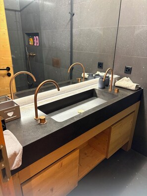 Un baño moderno con encimera negra, dos grifos de cobre, un espejo grande y armarios de madera.