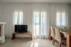 A living room with a flat-screen TV on a wooden bench, white curtains, a dining table with wicker chairs, and a tiled floor. - Kriolithos Houses (Paros)