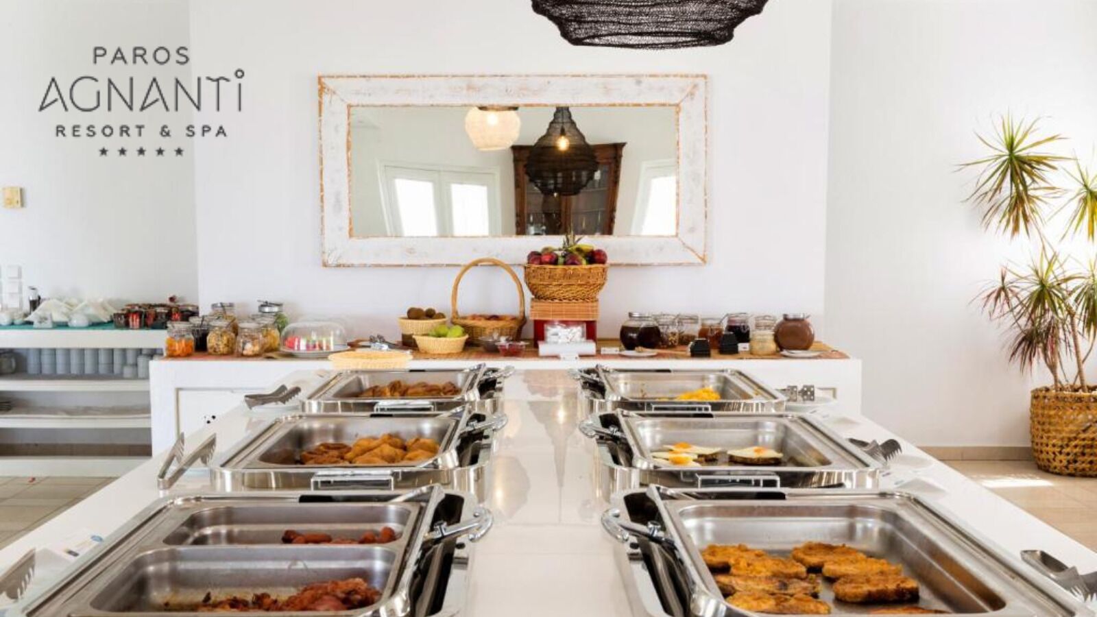 A buffet setup with various food items in metal containers, a mirror on the wall, and a sign for 'PAROS AGNANTI RESORT & SPA'.