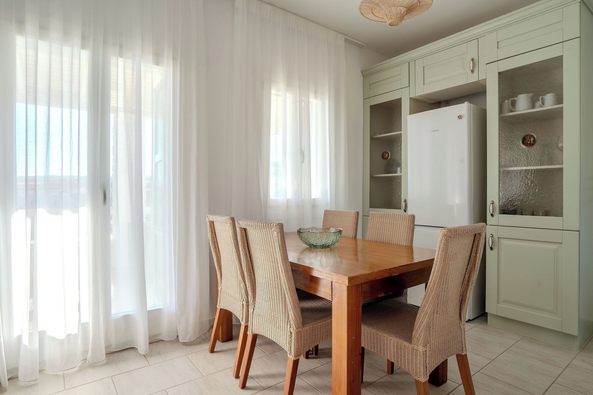 Un coin repas avec une table en bois et des chaises en osier, un réfrigérateur blanc et un meuble avec des portes en verre.