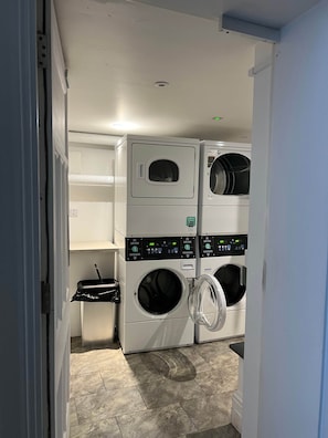 A laundry room with a stacked washer and dryer unit, a trash bin, and a door leading to another room. - Cozy city centre apartment | LGI and shops (Leeds)