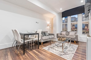 A modern living room with a dining area, grey sofas, a glass coffee table, and a view of the city through large windows. - Cozy city centre apartment | LGI and shops (Leeds)