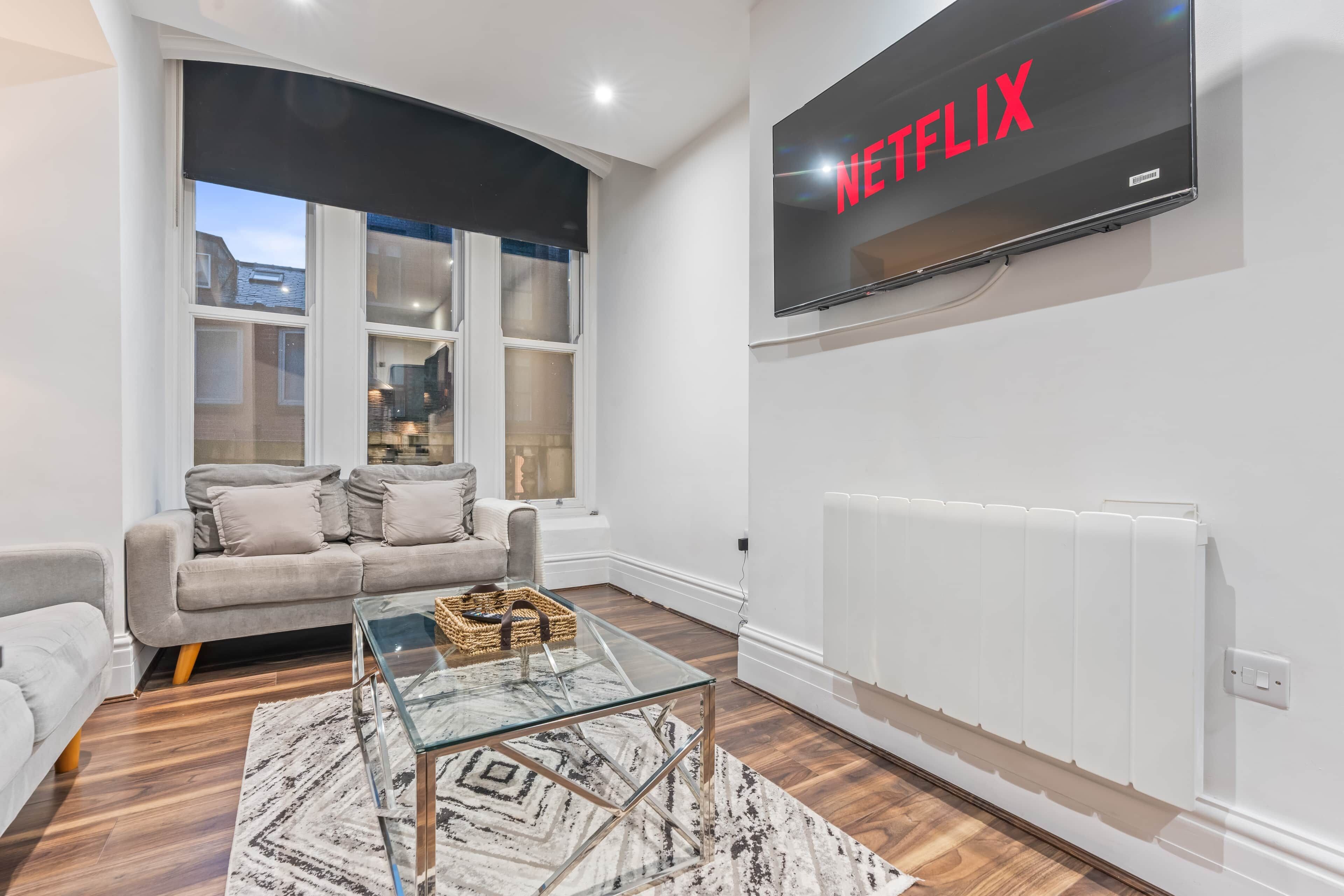 A modern living room with a grey sofa, a glass coffee table, and a large flat-screen TV mounted on the wall displaying 'NETFLIX'.