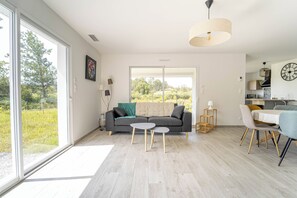 A modern living room with a sofa, two small tables, and a dining area with chairs. There is a large window providing a view of greenery outside. - Comfortable house between forest and ocean (Mimizan)