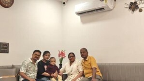 Une famille est assise sur un canapé dans un salon, où l’on peut voir un ventilateur de plafond, un appareil de climatisation et un objet décoratif fixé au mur.