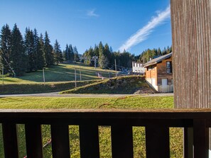 A wooden deck overlooking a grassy area with a building and a ski lift in the background. - Cozy studio for 3, piste view (Morillon Village)