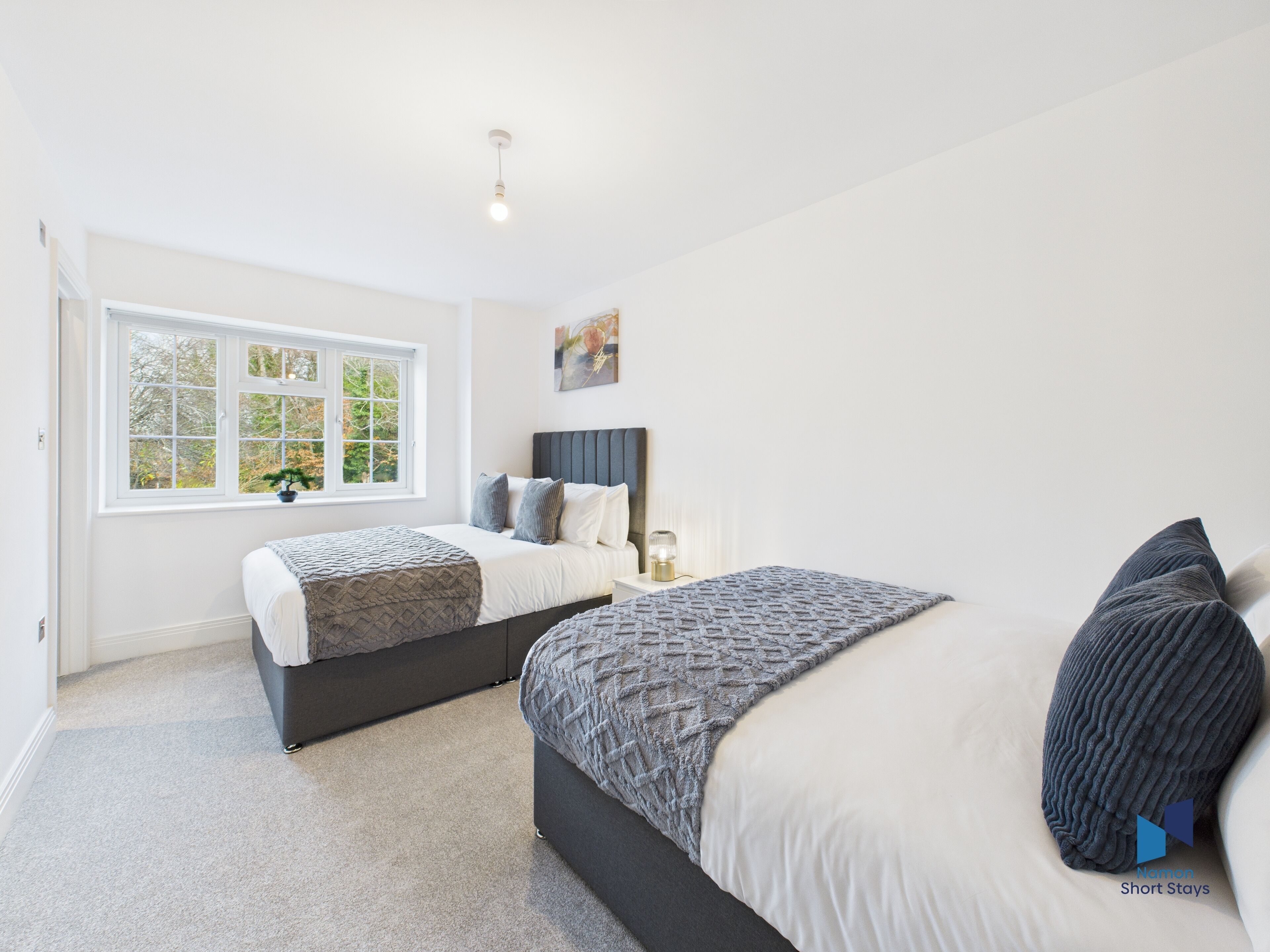 A bedroom with two beds, a window with a view of trees, a bedside table with a lamp, and a painting on the wall.