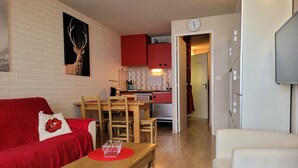 A cozy living room with a red sofa, a wooden coffee table, and a white armchair. A kitchen is visible in the background with red cabinets and a clock on the wall. - Bright studio 4 pers, balcony, close to slopes Pra-Loup 1600 (Pra-Loup)