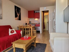 Ein gemütliches Wohnzimmer mit einem roten Sofa, einem Holztisch und einem weißen Sessel. Im Hintergrund ist eine Küche zu sehen, mit roten Schränken und einer Uhr an der Wand.