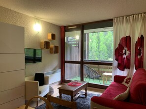 Ein gemütliches Wohnzimmer mit einer Couch, einem kleinen Tisch, einem an der Wand montierten Fernseher und einem Fenster mit Vorhängen.