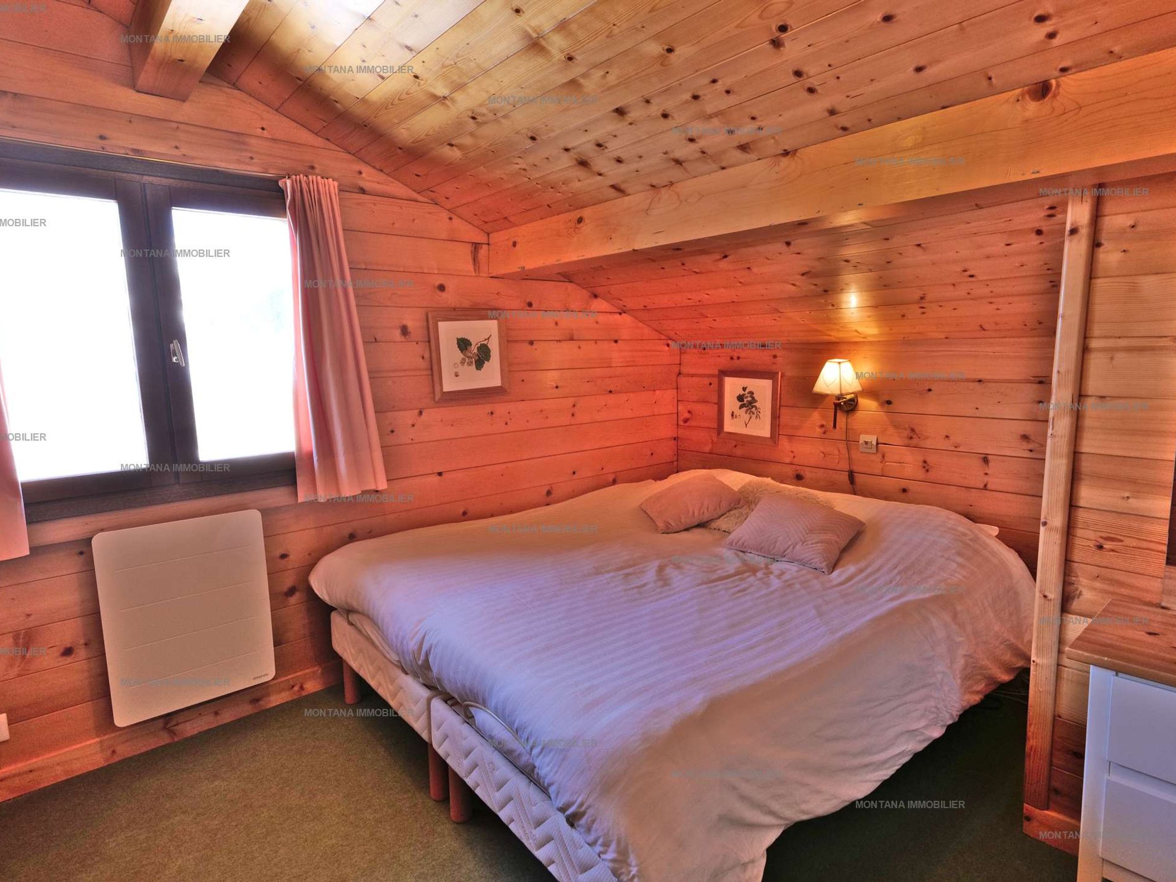 A cozy wooden cabin bedroom with a bed, a window with curtains, and a wall-mounted lamp.