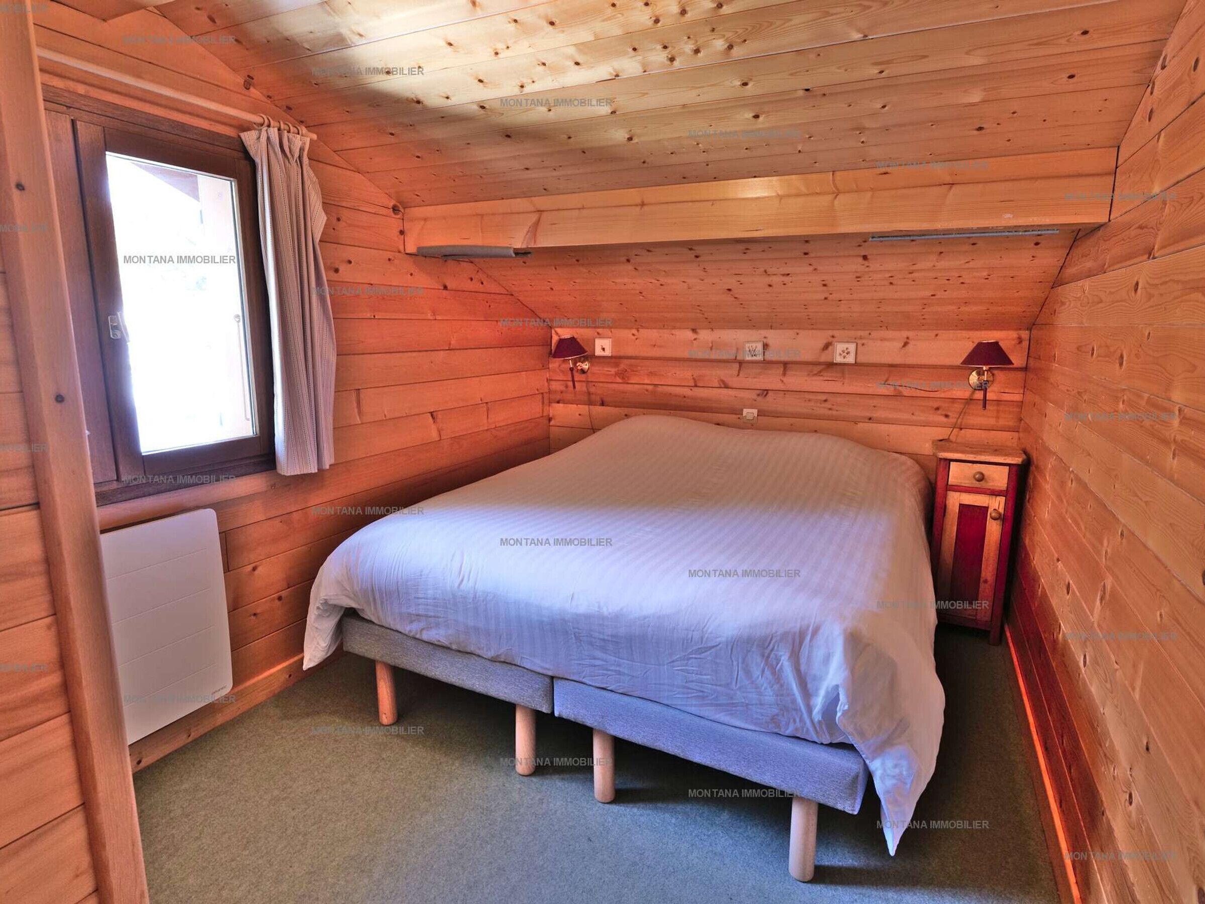 Une chambre dans une cabane en bois, avec un lit, une fenêtre avec des rideaux, une table de chevet et une lampe fixée au mur.