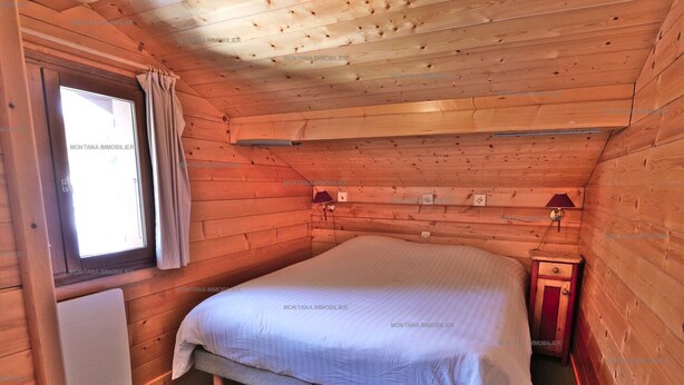 Ein Schlafzimmer einer Holzhütte mit einem Bett, einem Fenster mit Vorhängen, einem Nachttisch und einer Wandlampe.