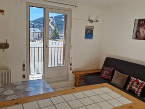 A room with a view of a town through a sliding door, a small wooden table, a sofa with plaid cushions, and a poster on the wall. - Studio Cabin in Les Angles center, practical with sleeping area (Les Angles)