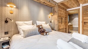 Un dormitorio acogedor con cabecera de madera, una cama grande con ropa de cama blanca, un oso de peluche en la cama y un armario de madera con espejo.