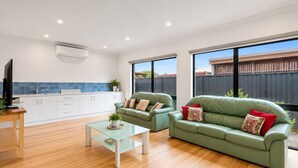 A modern living room with a glass coffee table, two green sofas with patterned cushions, a flat-screen TV, and a kitchen area with white cabinets and a blue backsplash. - Mercer on Port - Your Perfect Portarlington Getaway (Portarlington)