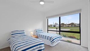 A bedroom with two beds, a ceiling fan, and a large window with a view of a residential area. - Mercer on Port - Your Perfect Portarlington Getaway (Portarlington)