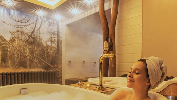 A person relaxing in a bathtub with a towel on their head, a wooden bench, and a view of trees through the window. - Hotel Franz - Rejvíz (Zlaté Hory)