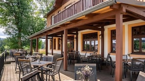 An outdoor dining area with wooden tables and chairs, a wooden deck, and a building with large windows and a balcony. - Hotel Franz - Rejvíz (Zlaté Hory)
