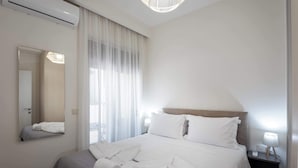 A hotel room with a bed, a bedside table with a lamp, a mirror, and a window with sheer curtains. - Harma City Living - Street View Standard Apartment (Chania)