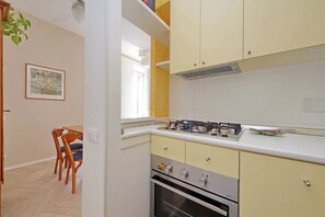 Eine Küche mit Gasherd, Backofen und gelben Schränken. Ein Esstisch mit Stühlen und ein gerahmtes Bild an der Wand sind ebenfalls vorhanden.