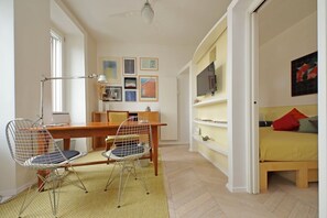 Ein schlichter Raum mit einem Holztisch, zwei Drahtstühlen, einem Schreibtisch mit Lampe, einem Bett mit gelber Bettdecke und einem Fernseher auf einem Regal.