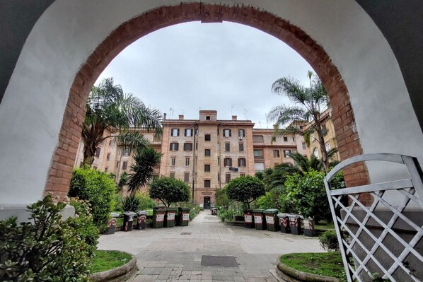 An arched entrance leading to a building with a central courtyard, surrounded by greenery and trash bins. - CASA SUL PORTO ANTICO (Roma)