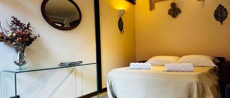 A hotel room with a bed, a glass desk, a vase with flowers, a mirror, and wall-mounted lights.
