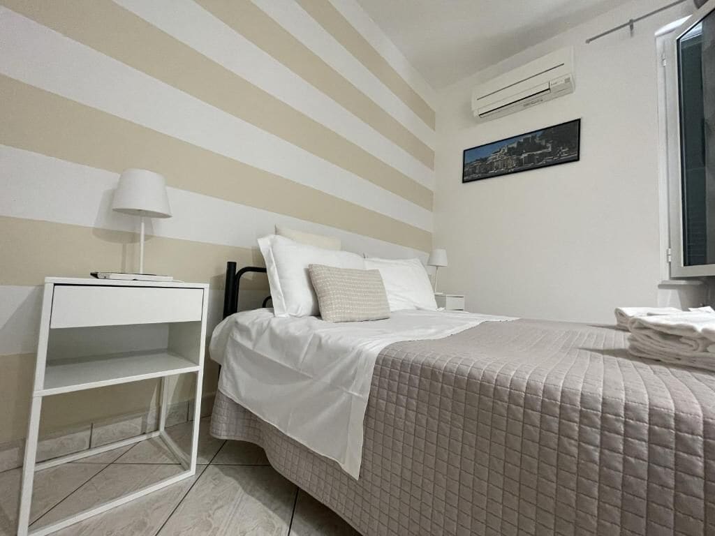 A hotel room with a bed, a white nightstand, a wall-mounted air conditioner, and a window with a view.