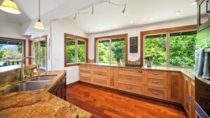 A modern kitchen with granite countertops, wooden cabinets, and large windows offering a view of greenery. - Ohana Kai, Luxe Princeville home w/ AC (Princeville)