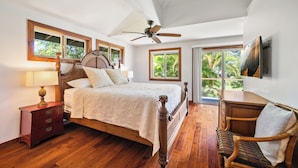 A bedroom with a wooden bed, a nightstand, a chair, a ceiling fan, and a large window with a view of greenery. - Ohana Kai, Luxe Princeville home w/ AC (Princeville)