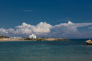 A coastal scene with a white building near the shore, a rocky outcrop, and a clear blue sky with clouds. - Villa Melinda: Comfort & Privacy Near the Beach (Protaras)