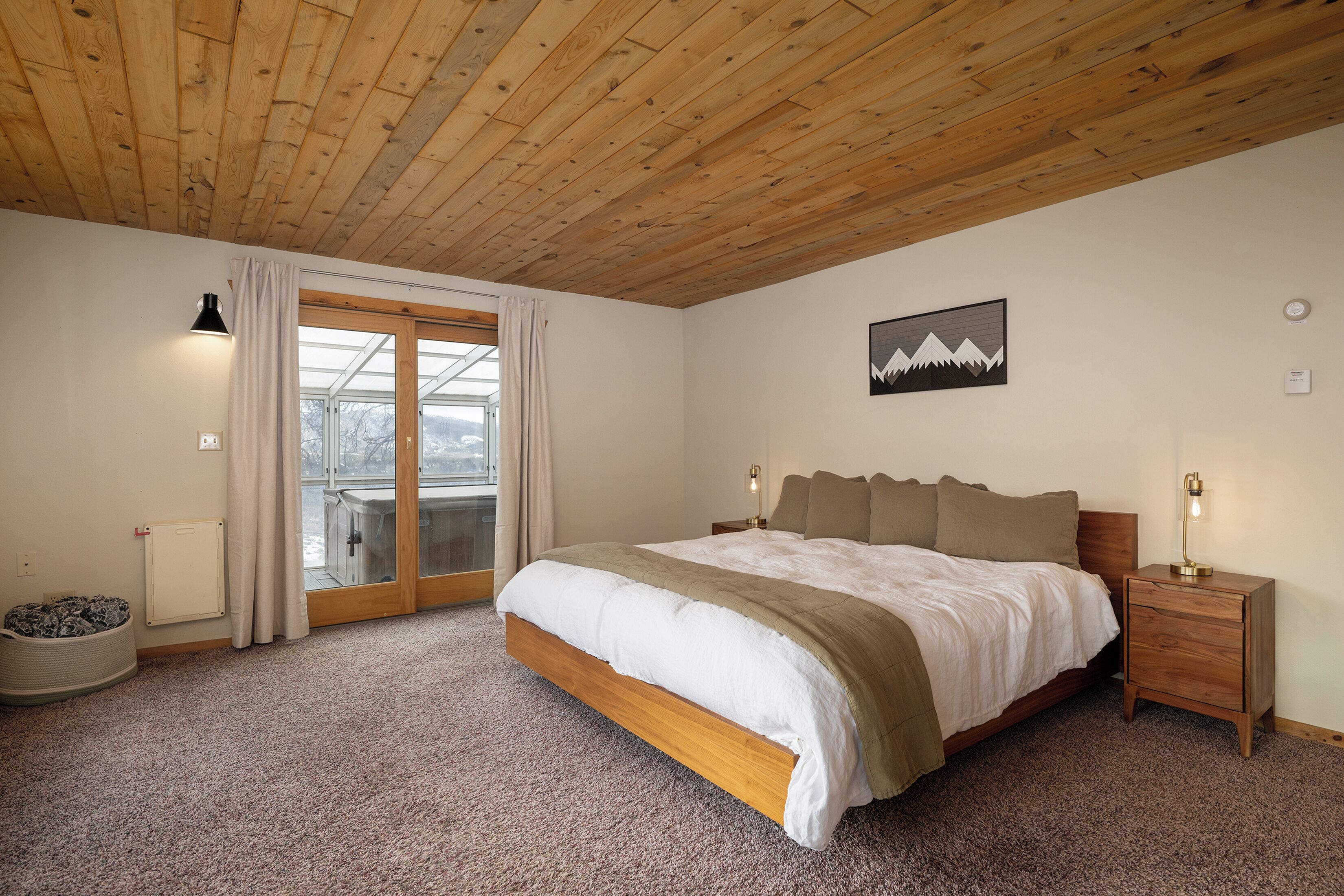 Una camera da letto con soffitto in legno, un letto grande, un comodino, un termosifone, una finestra con tende e un'opera d'arte raffigurante una montagna al muro.