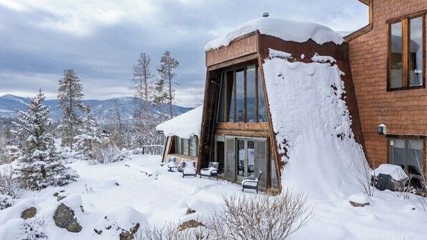 Ett modernt hus med stora glasfönster, en fasad av tegel och ett snötäckt landskap med berg i bakgrunden.