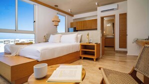 Une chambre à coucher moderne avec un grand lit, des tables de chevet en bois, un coin lecture avec un livre et une tasse, et une vue sur la ville à travers de grandes fenêtres.