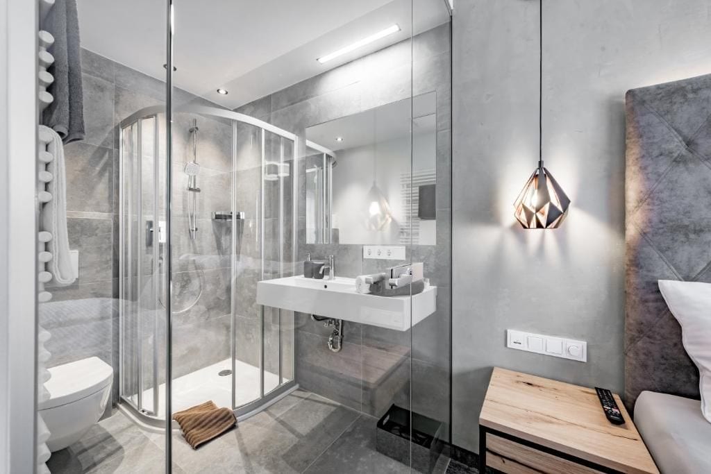 A modern bathroom with a glass-enclosed shower, a white sink, and a bedside table with a remote control.