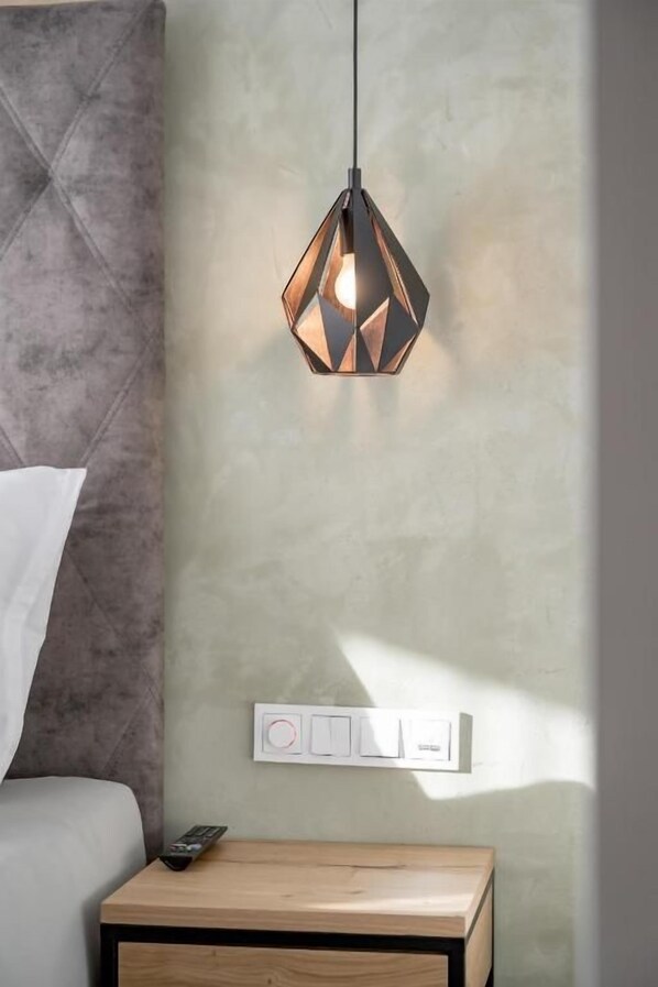 A modern bedroom with a geometric pendant light, a bedside table with a remote control, and a wall-mounted light switch.