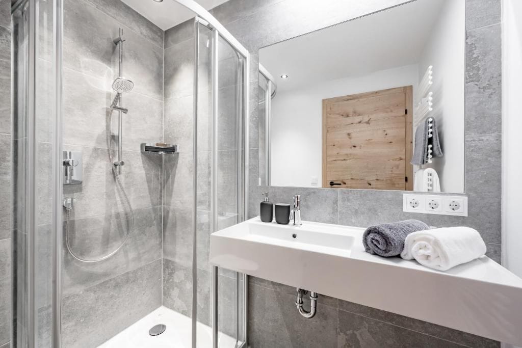 A modern bathroom with a glass-enclosed shower, a white sink, and a wooden door.